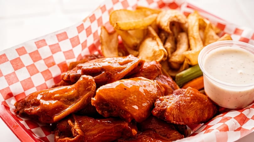 Basket of glossy BBQ chicken wings, potato wedges, celery, carrots, and ranch dressing.