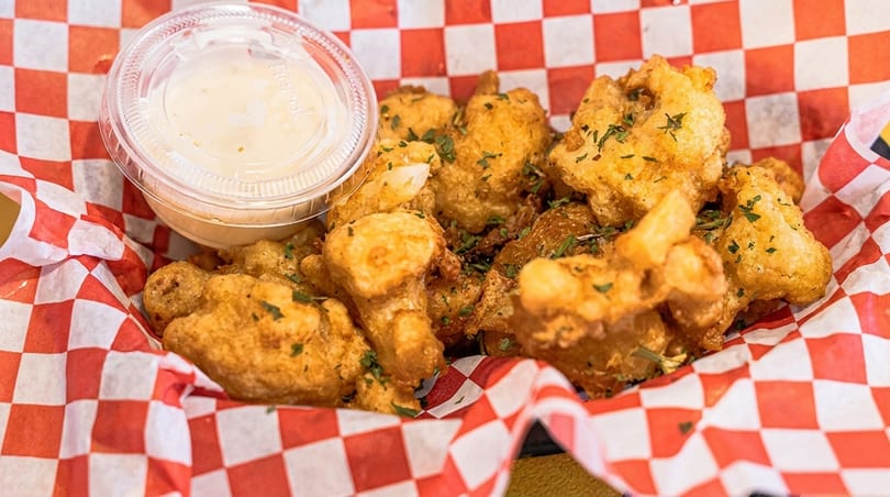 Golden fried cheese curds with parsley flakes and a side of dipping sauce in a red and white checkered basket.