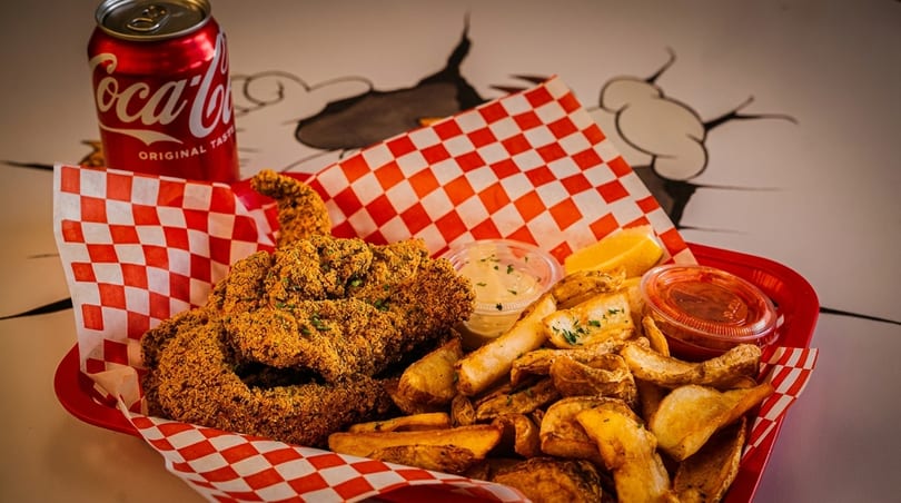 Fried fish and potato wedges in a red basket with sauces and a Coca-Cola can.