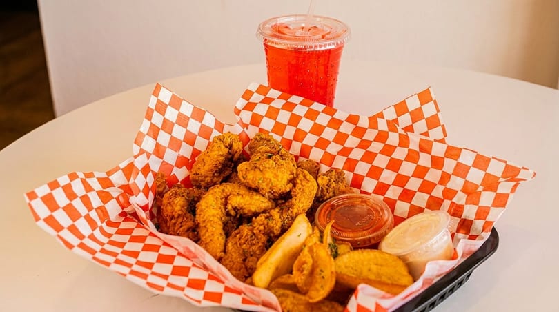 Fried chicken and potato wedges in a basket with red checkered paper, alongside a red drink.