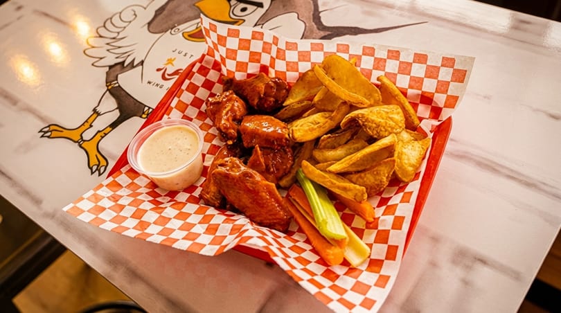 A red basket with checkered paper holds buffalo wings, potato wedges, celery, carrots, and ranch dressing.