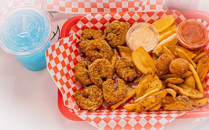 Fried shrimp, potato wedges, and hush puppies in a red basket with a blue drink.